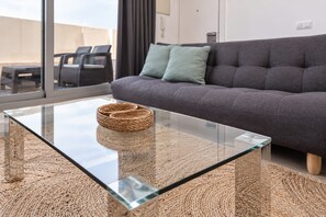 Een moderne woonkamer met een glazen bijzettafeltje, een geweven vloerkleed, een grijze bank en uitzicht door grote ramen.