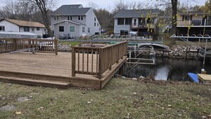 Une terrasse en bois avec une rambarde en métal, un petit quai s’avançant dans l’eau, et des maisons en arrière-plan.