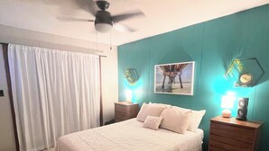 Une chambre à coucher avec un lit, un ventilateur de plafond, une table de chevet avec une lampe et un tableau accroché au mur.