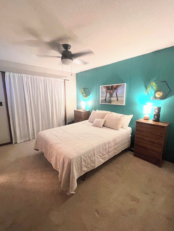 Un dormitorio con cama, ventilador de techo, mesita de noche con lámpara y un cuadro colgado en la pared.