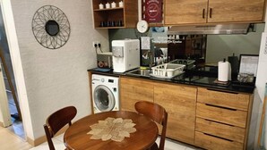 A compact kitchen with wooden cabinets, a built-in washing machine, a coffee maker, and a dining area with a table and chairs. - Timurbay Residence Resort Style (Kuantan)