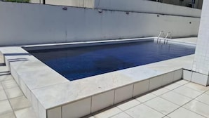 Una piscina rectangular con un borde de baldosas, una escalera y una pared blanca al fondo.