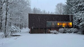A modern, dark-colored building with a large window and a visible entrance. Snow covers the ground and trees, indicating a winter setting. - Contemporary Hudson Valley home (Staatsburg)