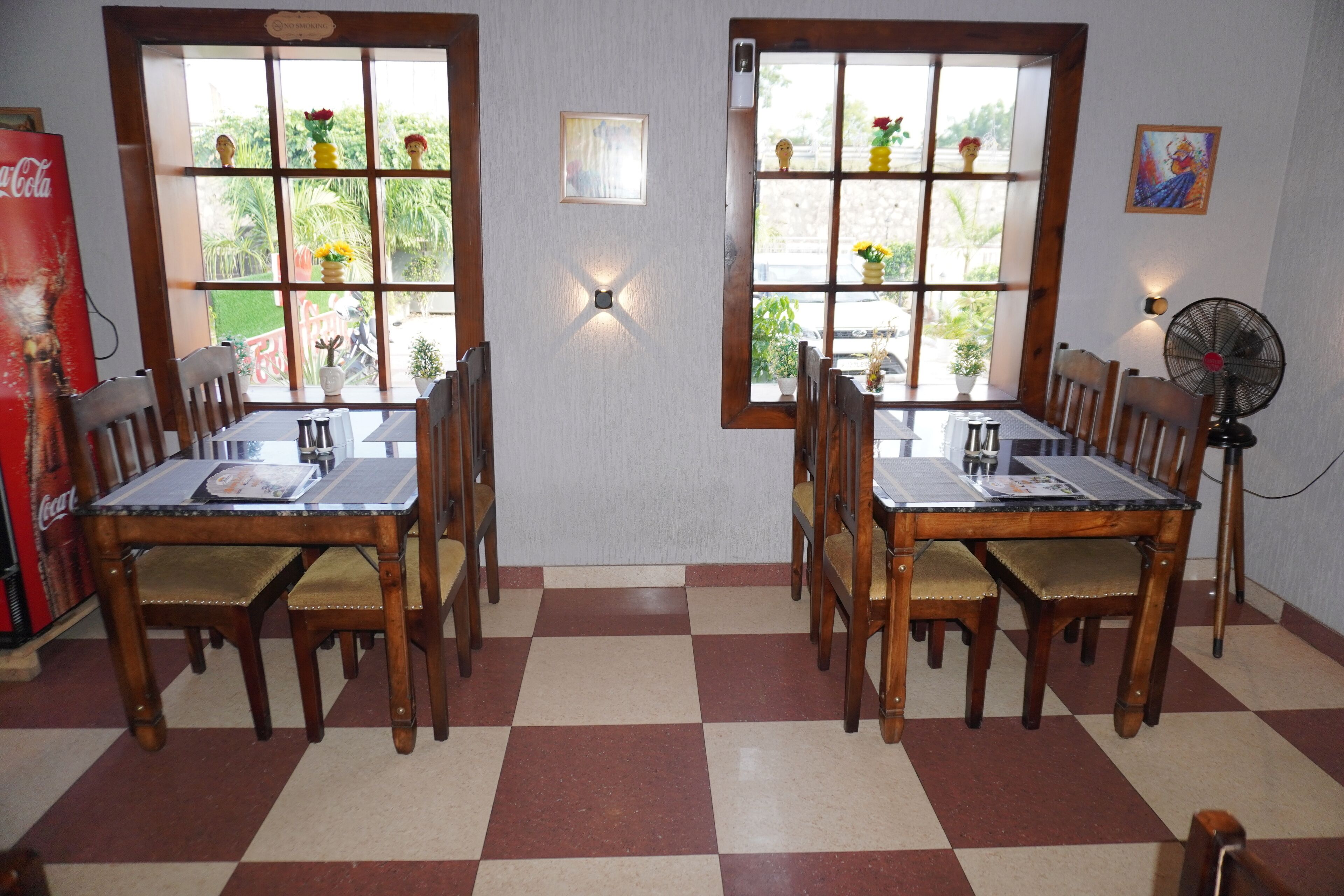 A dining area with wooden tables and chairs, large windows with view of greenery, and a fan on the right.