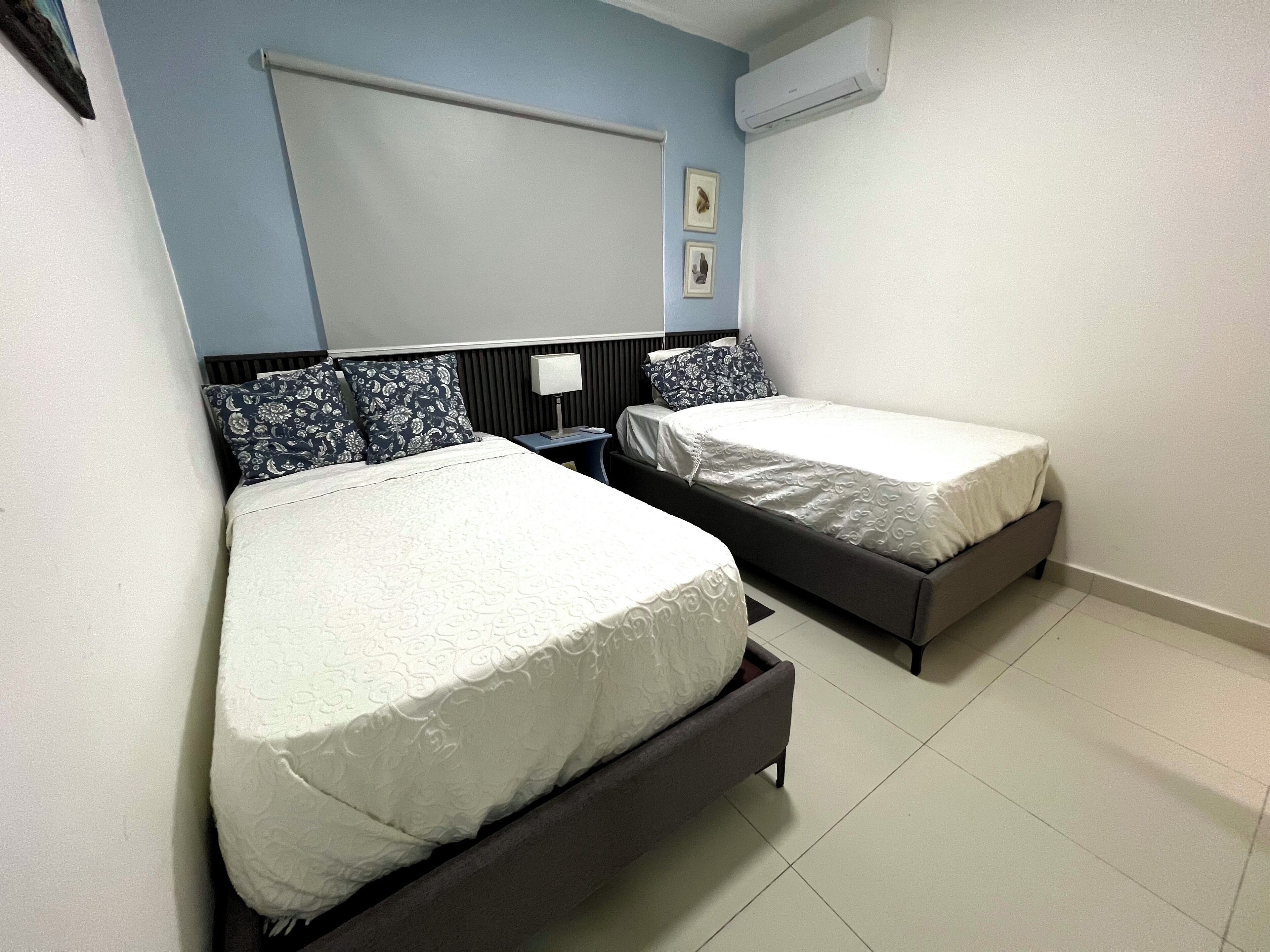 A room with two beds, a nightstand, a wall-mounted air conditioner, and a window with blinds.