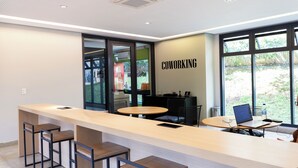 Un espacio de coworking moderno con una larga barra, varios taburetes, una mesa redonda con una computadora portátil y grandes ventanales que ofrecen vistas a zonas verdes.