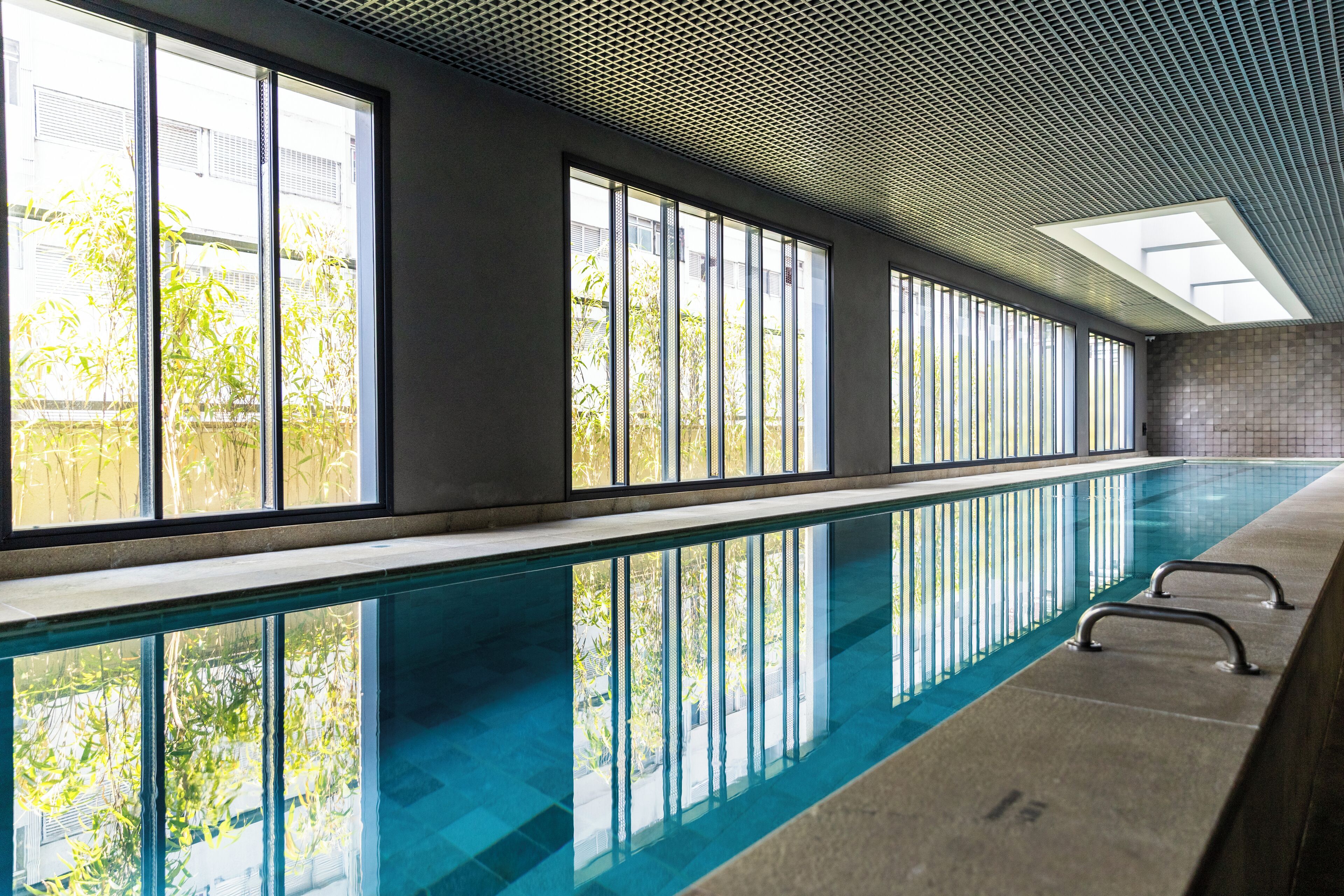 Une piscine intérieure avec des parois en verre transparent, un sol carrelé et un plafond orné d’un motif ajouré.
