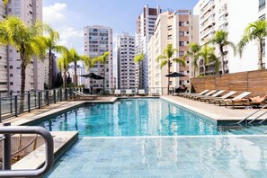 A swimming pool with lounge chairs and palm trees, surrounded by tall residential buildings. - Piscina- academia- AC - Brazilian Corner (São Paulo)