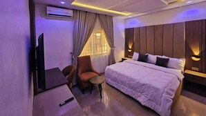 Ein modernes Hotelzimmer mit einem großen Bett, einem Schreibtisch und einem Sessel. Ein Fernseher ist an der Wand angebracht und ein Fenster mit Vorhängen vorhanden.