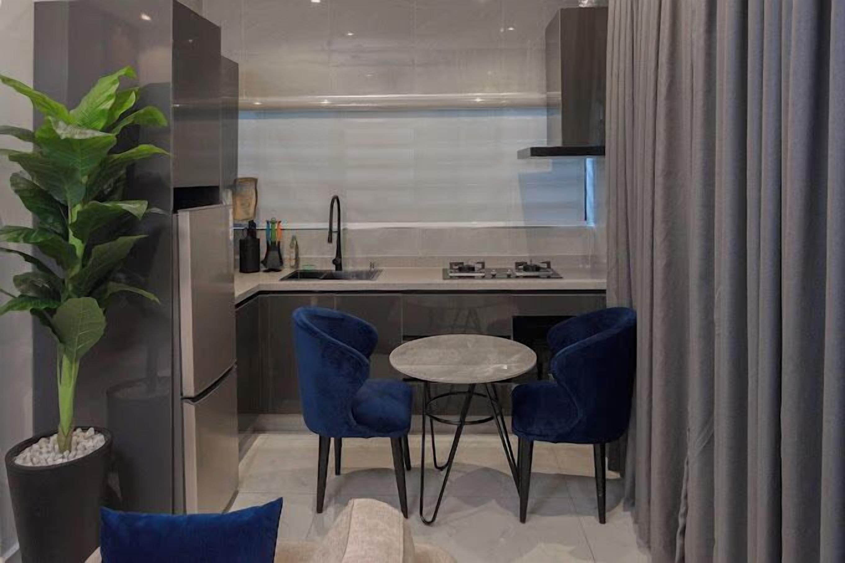 Uma cozinha compacta com design moderno, com frigorífico em aço inoxidável, uma pia e um pequeno espaço de refeições com duas cadeiras azuis e uma mesa redonda.