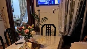 A dining area with a table covered in a floral tablecloth, a vase with flowers, and a tray with a house model. A television displaying 'STILL STANDING' is mounted on the wall. - Ρετιρέ μεγάλο 3 δωμάτια.καθιστικό με βεράντα αγ. Κωσταντίνος με θέα το Πήλιο (Volos)