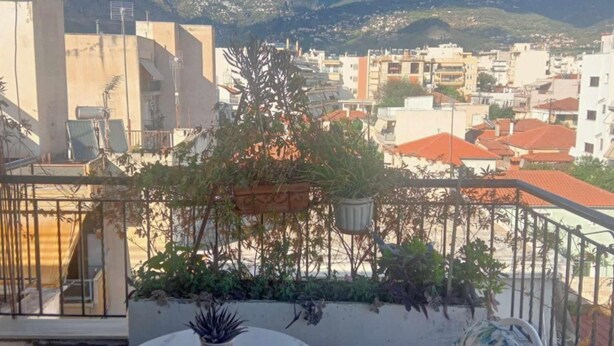 A terrace with a table and chairs, potted plants, and a view of a cityscape with mountains in the background. - Ρετιρέ μεγάλο 3 δωμάτια.καθιστικό με βεράντα αγ. Κωσταντίνος με θέα το Πήλιο (Volos)