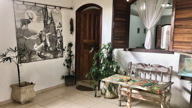 A room with a wooden door, a large painting, a potted plant, a table with a floral tablecloth, and a mirror with a curtain. - LARGE PRIVATE SUITE WITH BALCONY AND CLOSE TO THE HISTORIC CENTER. PET FRIENDLY  (Caborê Paraty)