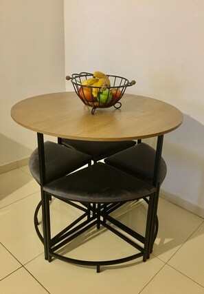 Een ronde tafel met een gevlochten mand vol fruit, twee zwarte stoelen met kussens, en een betegelde vloer.