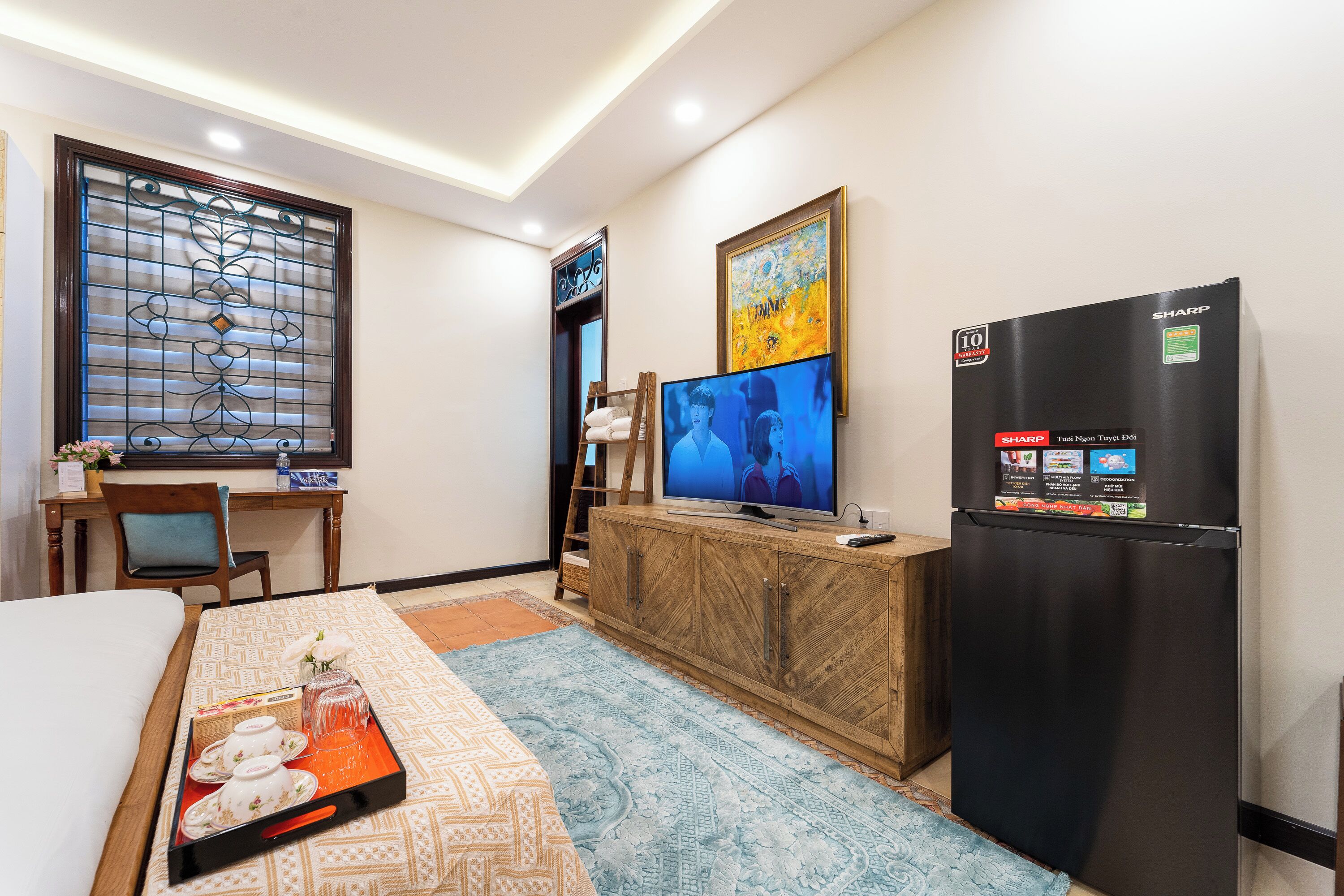 A hotel room with a bed, a wooden desk, a television, a refrigerator, and a patterned rug.