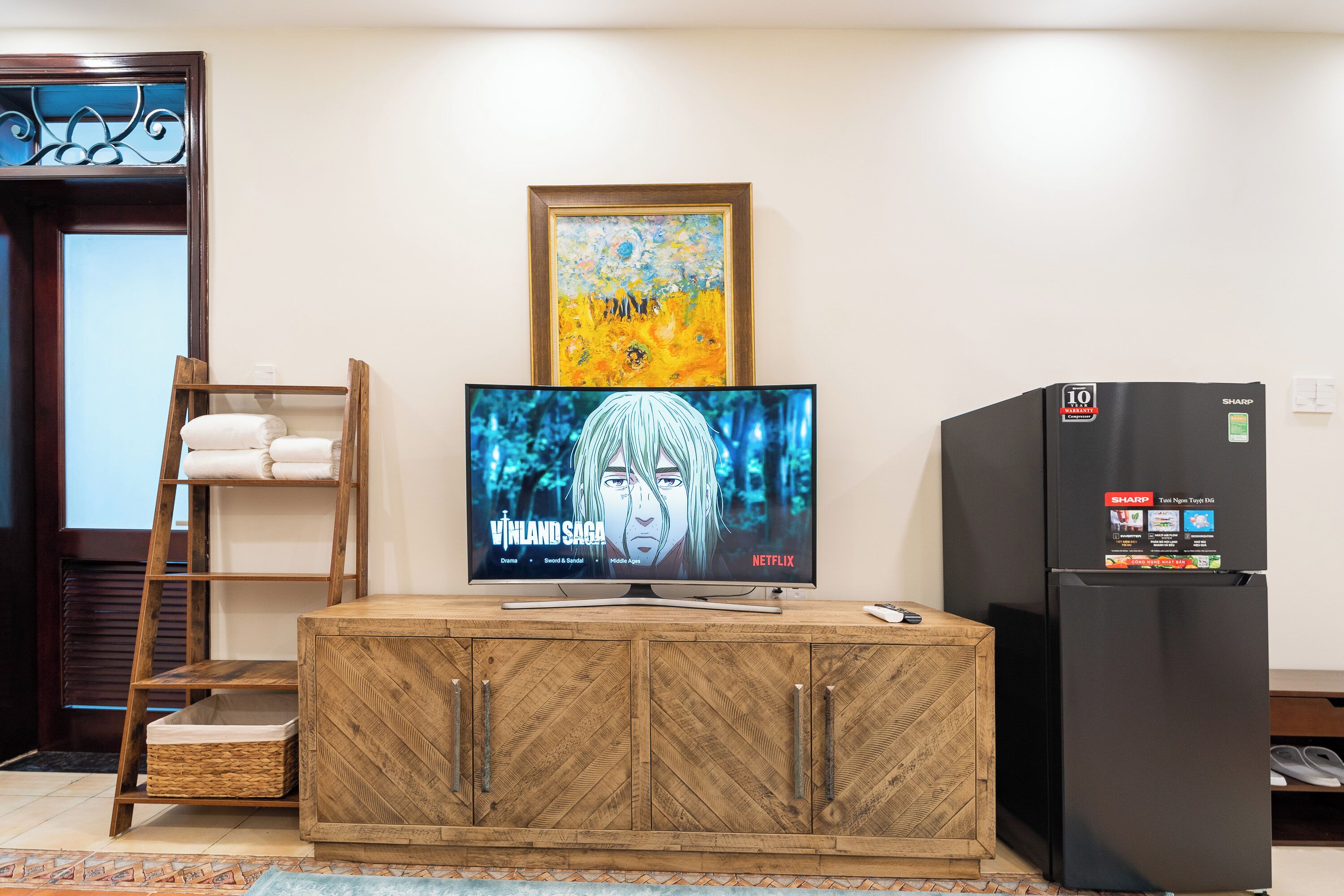 Un mueble de madera con un televisor de pantalla plana mostrando 'Vinland Saga' en Netflix, un refrigerador y una obra de arte enmarcada sobre el televisor.