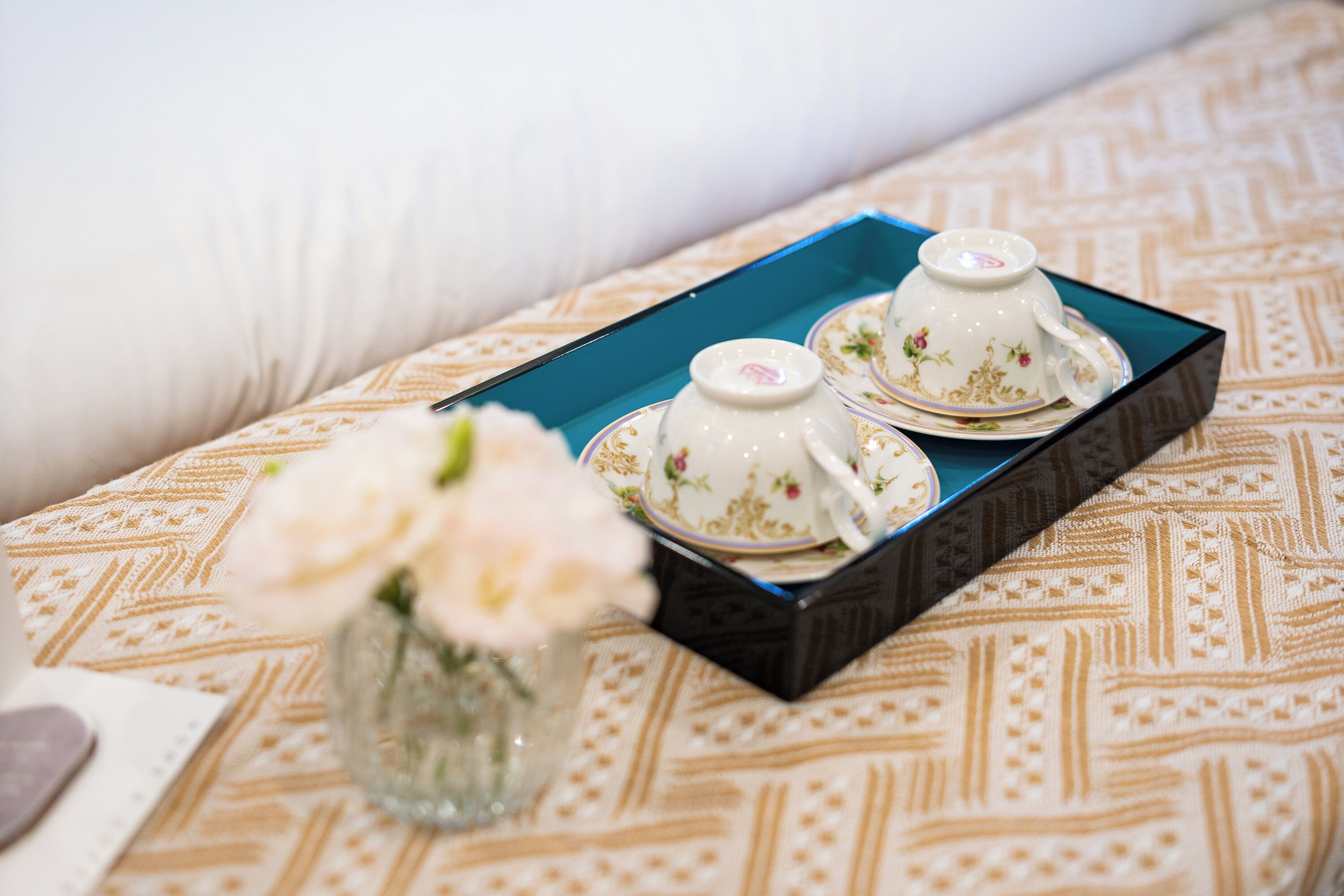 A tray with two teacups and saucers on a patterned tablecloth, accompanied by a vase with flowers.