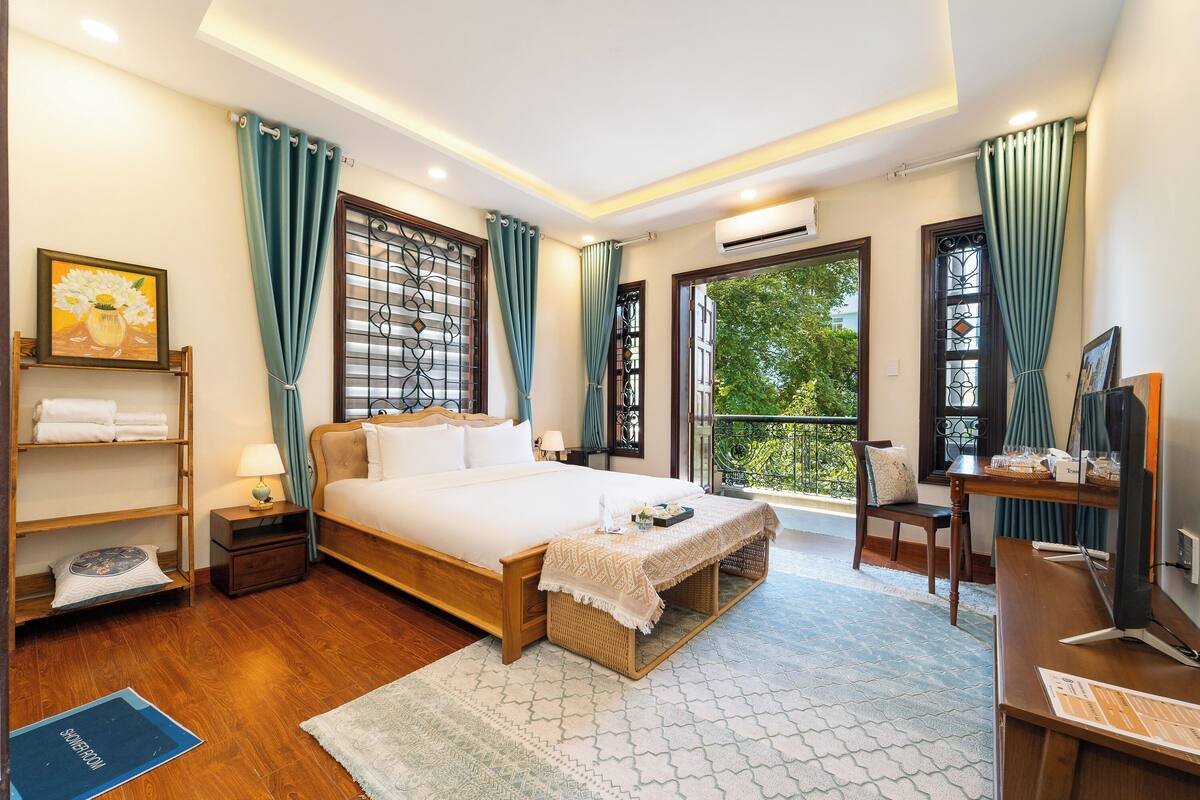 A well-lit bedroom with a large bed, a desk with a computer, and a view of greenery outside.