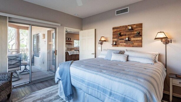 A bedroom with a large bed, a ceiling fan, a wooden wall decoration, and a balcony with a chair. - Walk to Fountain Park! Ground-Floor Condo w/ Pool, Spa & Private Patio (Fountain Hills)