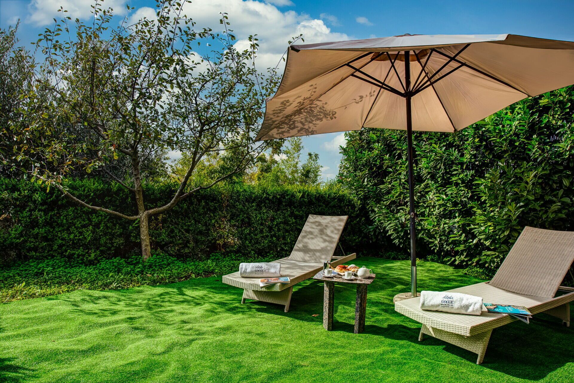 A garden with two wicker lounge chairs, a table with a newspaper and a teapot, and a large umbrella providing shade.