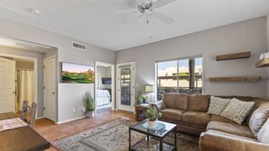 A living room with a brown leather sofa, a glass coffee table, and a patterned rug. There is a door leading to another room, a window with a view, and a ceiling fan. - Ground Floor Condo Near Plaza & Fountain Park w/ Pool, Hot Tub & Private Patio (Fountain Hills)
