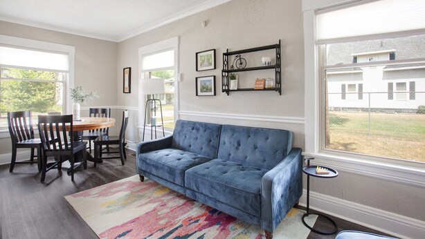 Een woonkamer met een blauwe bank, een eettafel, stoelen en een kleurrijk vloerkleed. Er hangen ingelijste foto's aan de muur en staat een klein bijzettafeltje.