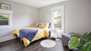 Een slaapkamer met een bed, nachtkastje, lamp en een raam met uitzicht op groen.