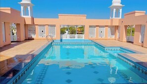 Una piscina amb un disseny de mosaic geomètric, envoltada d'un edifici beix amb detalls decoratius blancs i un cel blau clar al fons.