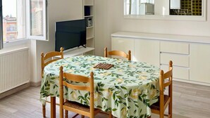 A dining area with a table set for four, a ceiling fan, a television, and a window with a view of buildings outside. - Magnificent 2p apartment in a villa with sea view, terrace & private parking. (Beaulieu-sur-Mer)