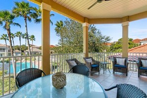 Un espacio exterior techado con mobiliario de mimbre y una mesa de vidrio, con vistas a una piscina y palmeras.