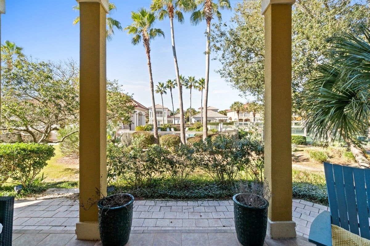 A covered outdoor area with a view of a residential complex, featuring palm trees and a paved walkway.