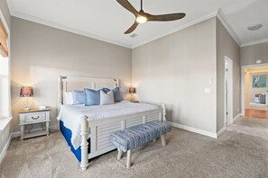 Une chambre à coucher comprenant un lit, des tables de chevet, un ventilateur de plafond et une porte donnant sur une autre pièce.