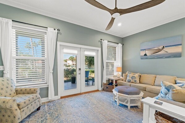 A room with a ceiling fan, a sofa, a patterned armchair, a small table with a lamp, and a painting of a boat on the wall.