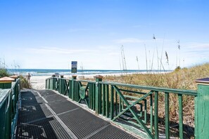 Ein Fußweg zum Strand mit einem Metallgitter, grünen Geländern und einem Wegweiser in der Ferne.