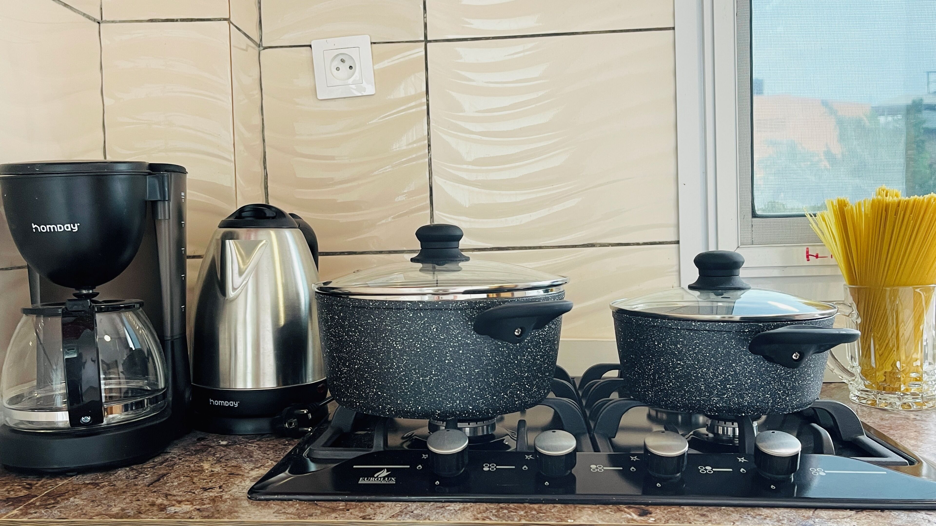 Una cocina con estufa, dos ollas, cafetera y un plato de espagueti.