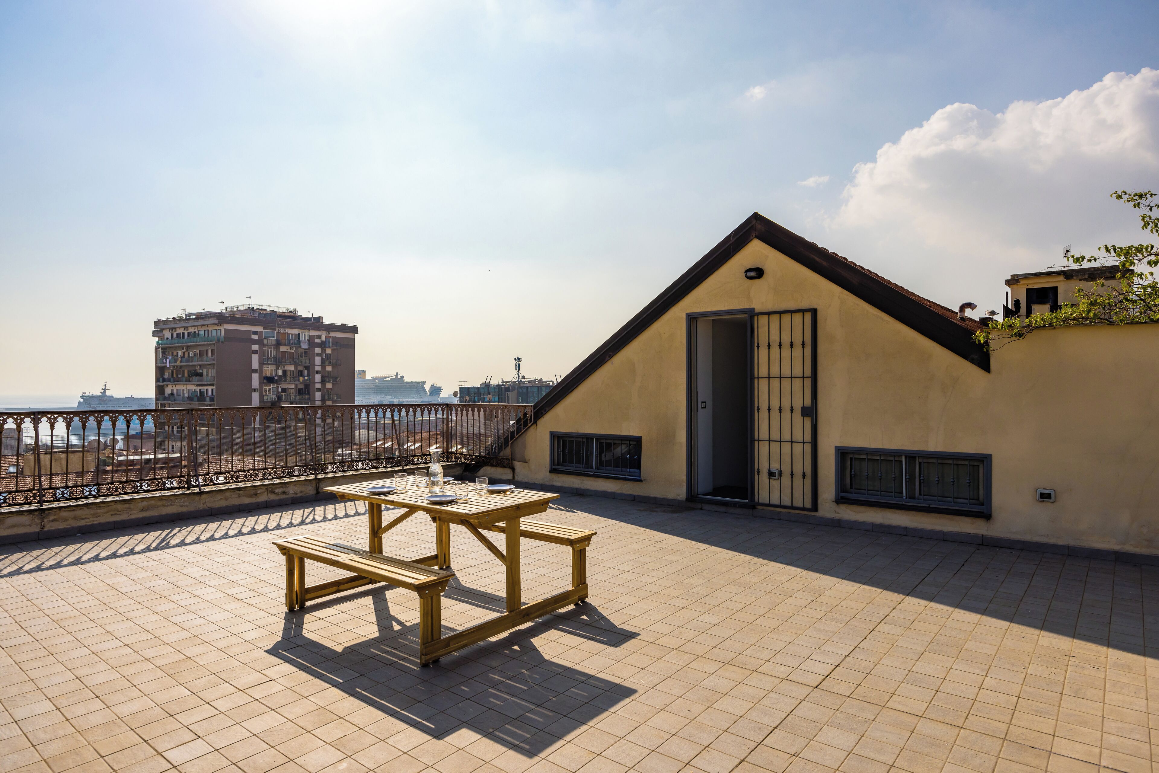 Una terraza en la azotea con una mesa y bancos de madera, vista al mar y un edificio con techo inclinado.