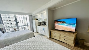 Habitación de hotel con dos camas, kitchenette y televisor mostrando una escena de playa.