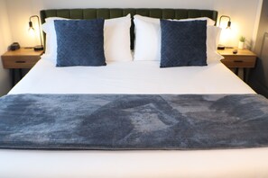 A bed with a white mattress, two pillows, a dark blue throw blanket, and a green upholstered headboard. There are two bedside tables with lamps and a small potted plant.