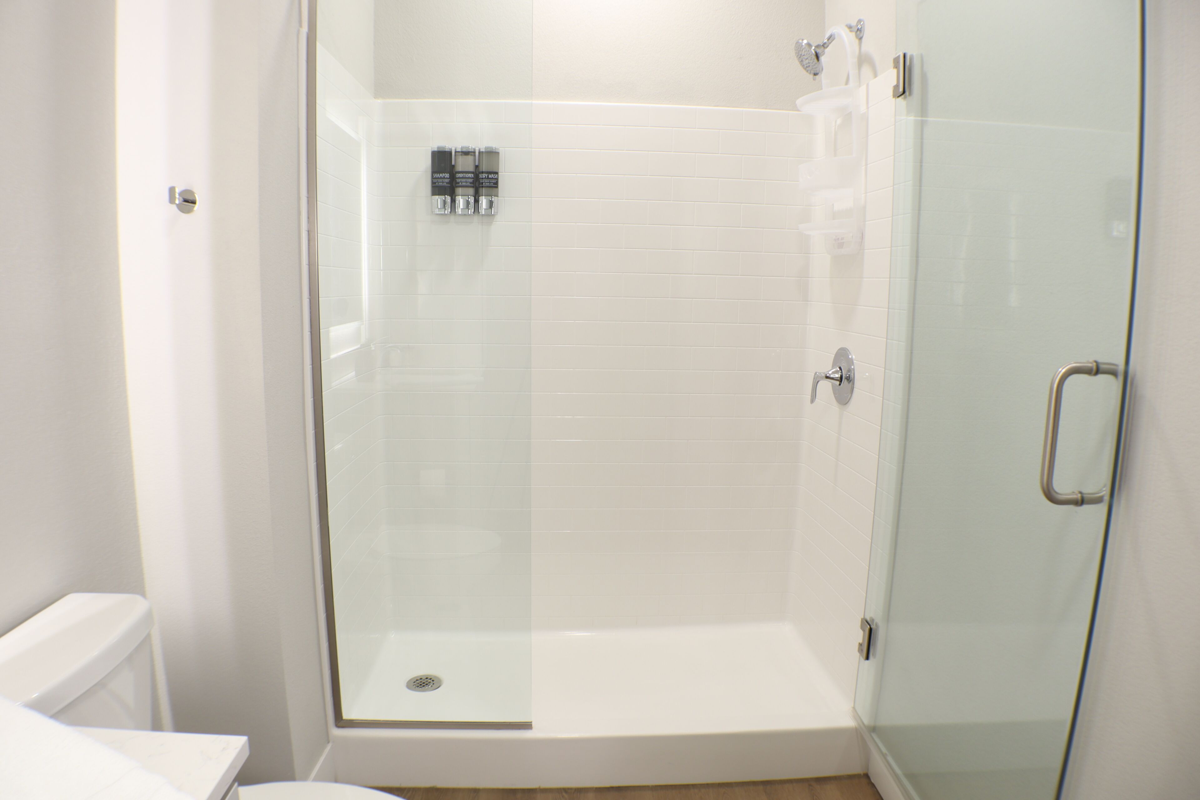 Une douche fermée par des parois de verre, avec un mur recouvert de carreaux blancs, un drain apparent et une barre d’appui en métal.