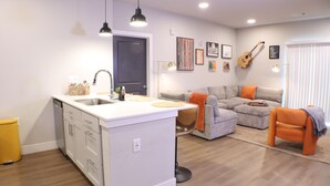 Una cocina moderna con una isla central, un área de comedor con un sofá y reposapiés, y una sala de estar con un sofá y una guitarra en la pared.
