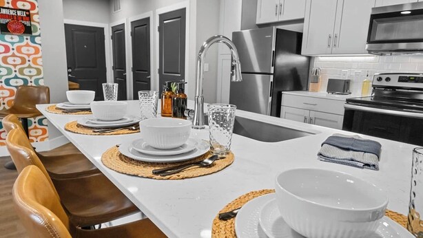 Una cocina moderna con encimeras blancas, electrodomésticos de acero inoxidable y una isla central con un lavamanos y asientos.