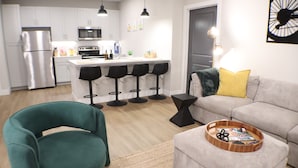 A modern kitchen with stainless steel appliances, a central island with bar stools, a grey sofa, and a wooden tray with magazines. - 2BR with king suites & pool near the Walmart NWA Championship. (Rogers)