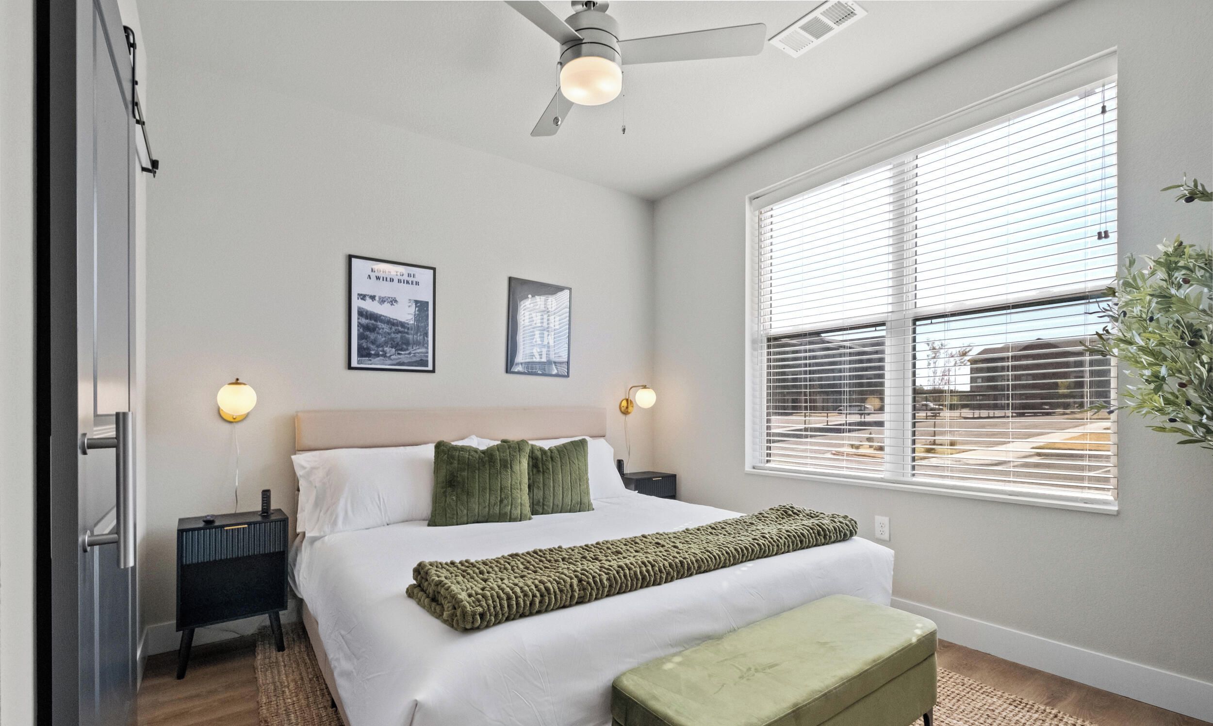 A bedroom with a bed, bedside tables, a ceiling fan, a window with blinds, and wall-mounted lights.