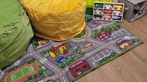 A child's play area with a yellow bean bag, a green bean bag, and a play mat featuring a cityscape with buildings like a school, firehouse, and bank. - Deluxe 2BR with king suites & pool near the Walmart NWA Championship. (Rogers)