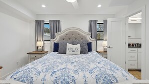 Une chambre à coucher avec un grand lit, une tête de lit capitonnée, des rideaux gris et une table de chevet avec une lampe.