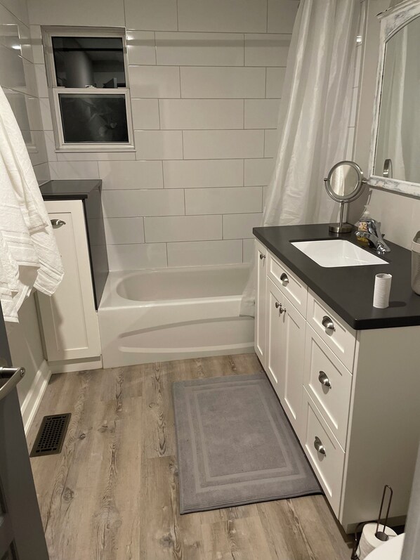 Une salle de bain avec un mur recouvert de carreaux blancs, une baignoire, un lavabo avec un miroir et un plancher en bois.