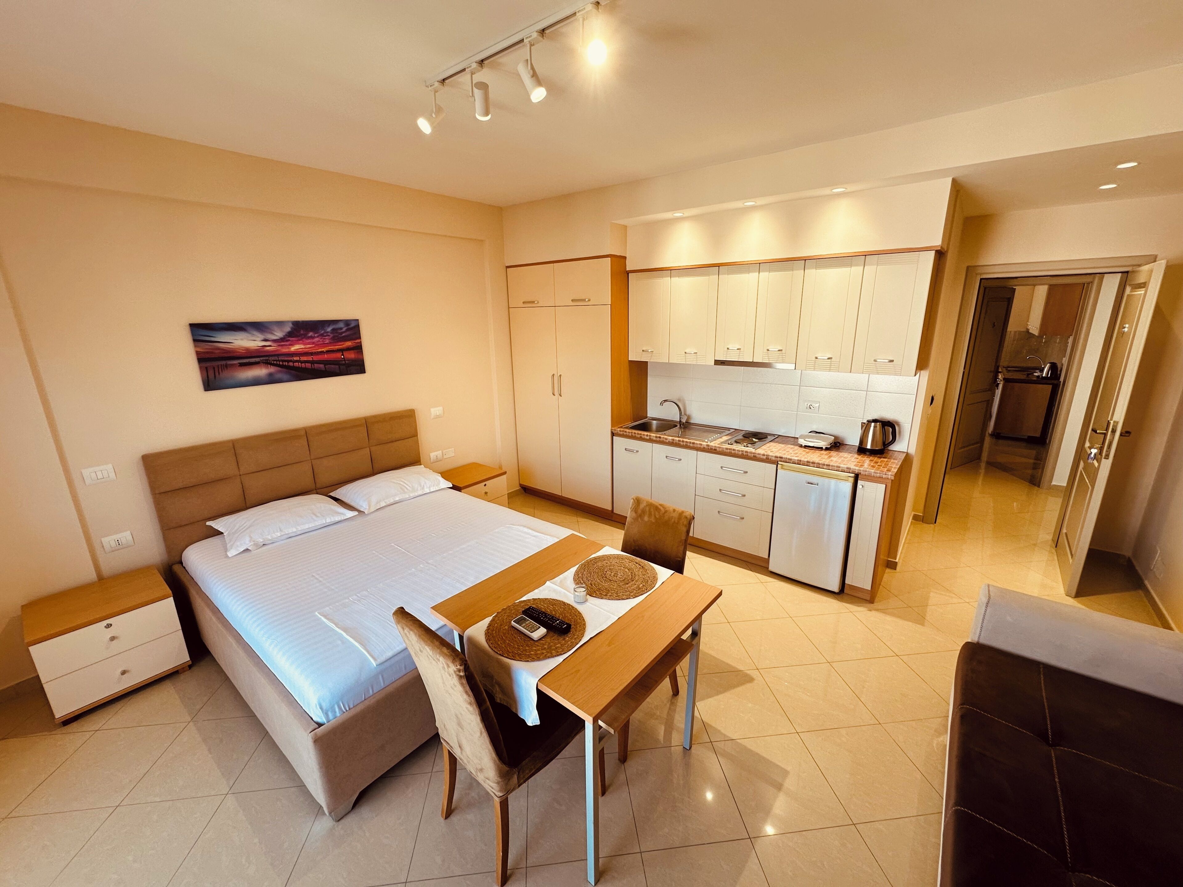A hotel room with a bed, bedside table, a small desk with a chair, a kitchenette, and a bathroom visible through an open door.