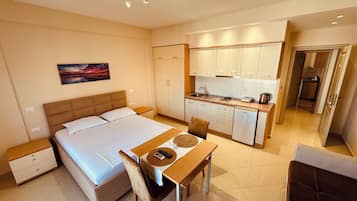 A hotel room with a bed, bedside table, a small desk with a chair, a kitchenette, and a bathroom visible through an open door.