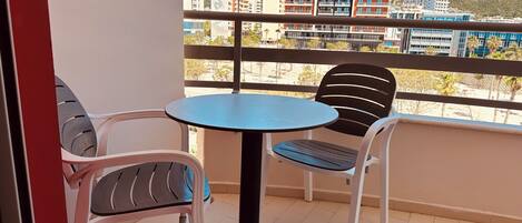 A balcony with a round table and two chairs, overlooking a cityscape with buildings and a hill.