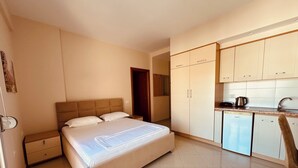 A bedroom with a bed, a nightstand, and a small desk with a chair. There is a kitchenette with a refrigerator, a sink, and a stove. - Thoma's K Studios (Vlorë)